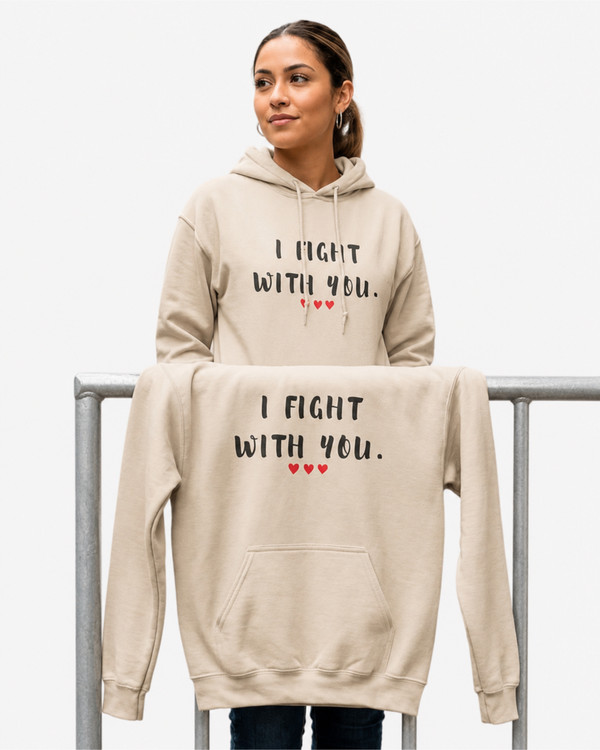 Woman wearing a sand-colored pullover hoodie featuring the supportive phrase “I Fight With You” in black distressed lettering with three red hearts beneath. A matching hoodie is displayed in the foreground. The hoodies feature a drawstring hood, front kangaroo pocket, and relaxed unisex fit. The design symbolizes encouragement, solidarity, strength, and standing together through life’s challenges.