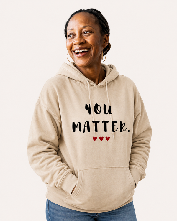 Woman wearing a sand-colored pullover hoodie featuring the uplifting phrase “You Matter” in distressed black lettering with three red hearts beneath. The hoodie is worn under a high-visibility safety vest and hard hat at a job site, symbolizing encouragement, self-worth, resilience, and support. The hoodie includes a drawstring hood, front kangaroo pocket, and relaxed unisex fit.