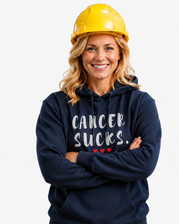 Woman wearing a navy blue pullover hoodie featuring the phrase “Cancer Sucks.” in distressed white lettering with three red hearts beneath. Standing confidently in an industrial workplace with a hard hat and forklift in the background, the image symbolizes strength, resilience, and support for cancer awareness. The hoodie combines comfort with a meaningful message of solidarity and hope.