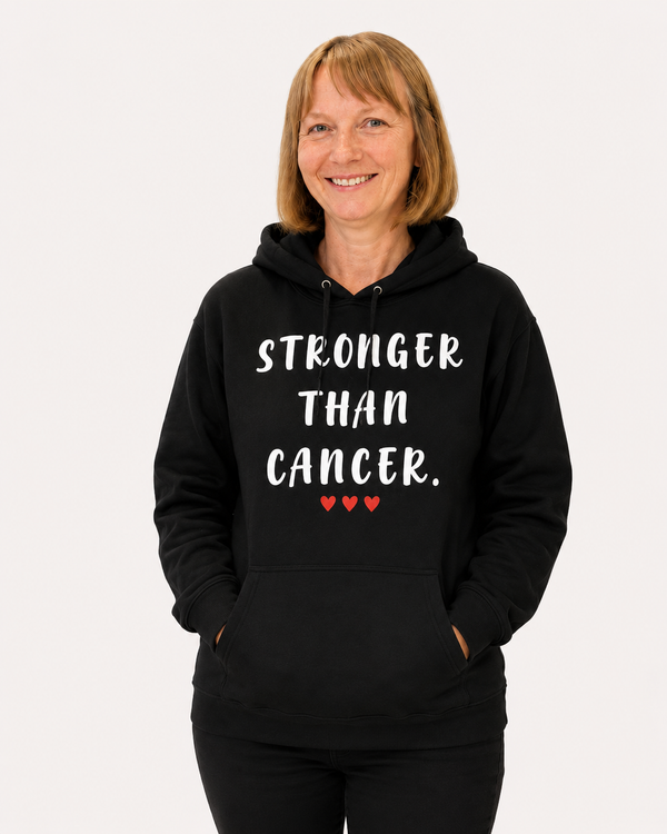 Smiling woman wearing a black hoodie with the inspirational message “STRONGER THAN CANCER.” printed in white lettering and three small red hearts underneath. She is standing indoors at a modern event or conference venue, with people blurred in the background. Several red metallic water bottles and a small teddy bear sit on the table in front of her. The hoodie design is clearly visible and serves as the focal point of the image, conveying strength, hope, and cancer awareness.