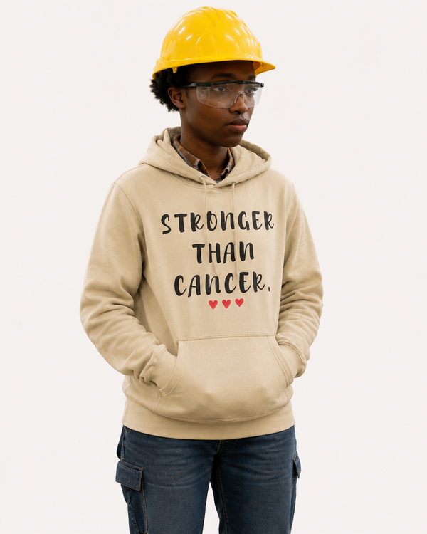 Person wearing a sand-colored pullover hoodie with the motivational message “STRONGER THAN CANCER.” printed in distressed black lettering across the chest, accented by three small red hearts beneath the text. The individual is standing in an industrial workshop environment and is wearing a yellow hard hat and safety glasses. Machinery and work equipment are visible in the softly blurred background. The image highlights strength, determination, resilience, and support for those affected by cancer while showcasing the hoodie design in a hardworking, professional setting.