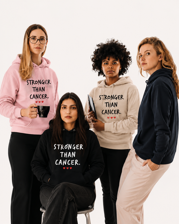 Four women gathered around a table in a bright, modern workspace. Three women are standing and one is seated behind a laptop. Two of the women are wearing hoodies featuring the inspirational message “STRONGER THAN CANCER.” in bold white lettering with small red hearts beneath the text. One hoodie is light pink and the other is sand beige. Coffee cups and camera equipment are arranged on the table, creating a collaborative and supportive atmosphere. The image conveys empowerment, unity, strength, and encouragement for those affected by cancer.