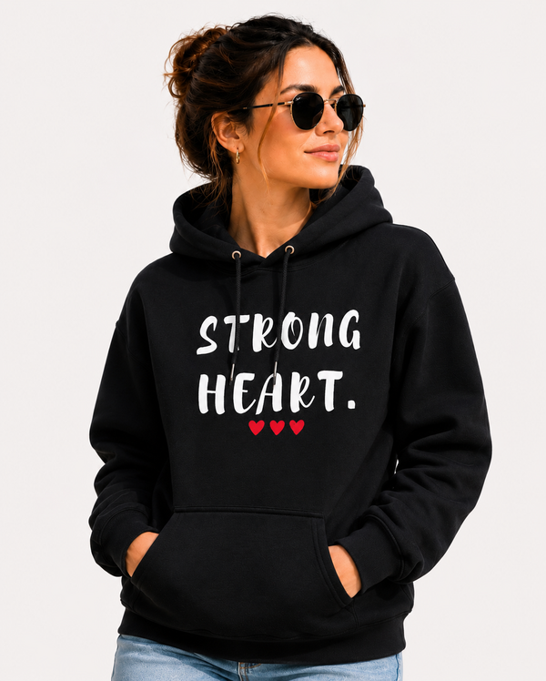Woman wearing a black hoodie with the empowering message “STRONG HEART.” printed in distressed white lettering and accented by three small red hearts beneath the text. She is standing along a scenic waterfront promenade, wearing sunglasses and smiling as she looks into the distance. Colorful buildings, people strolling, boats, and a marina create a vibrant coastal backdrop. The image conveys strength, resilience, confidence, and hope while showcasing the hoodie design in a stylish outdoor setting.