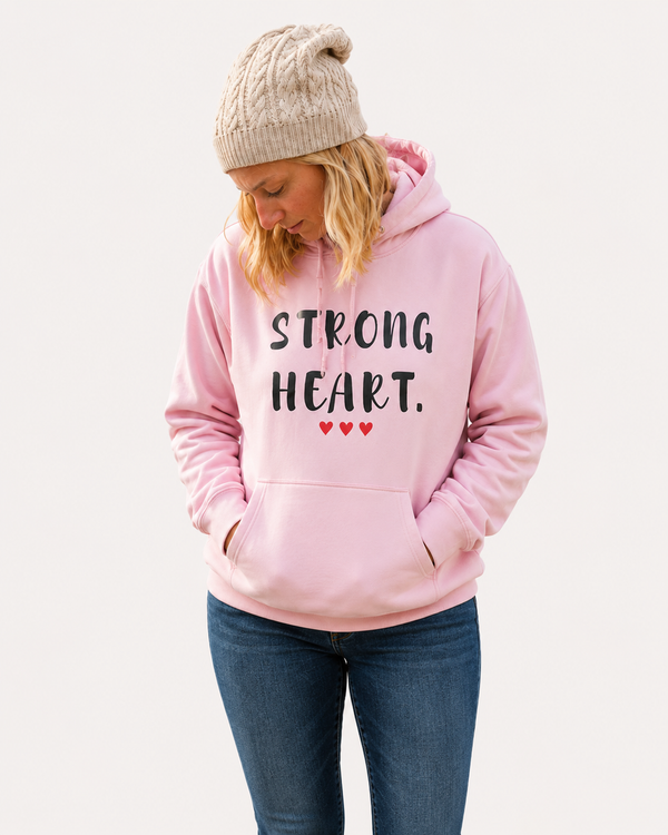 Woman wearing a light pink hoodie with the inspirational message “STRONG HEART.” printed in distressed black lettering and accented by three small red hearts beneath the text. She is crouching beside a crystal-clear mountain lake, gently touching the water while admiring its reflection. Snow-capped peaks, evergreen trees, and bright sunshine create a peaceful outdoor setting. The image conveys strength, resilience, inner peace, and a deep connection with nature while showcasing the hoodie design in an uplifting and scenic environment.