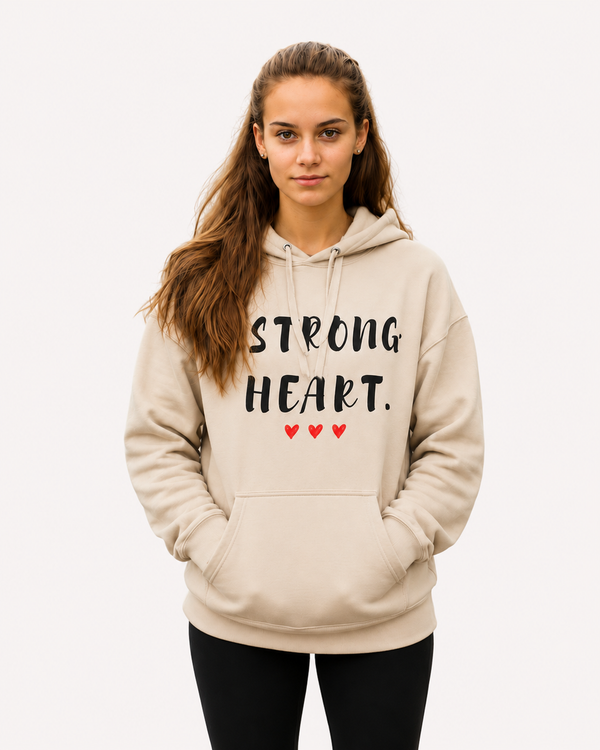 Young woman standing outdoors on a pathway wearing a sand-colored hoodie with the inspirational message “STRONG HEART.” printed in distressed black lettering and accented by three small red hearts beneath the text. She has an identification badge on a lanyard around her neck and her hands are tucked into the hoodie’s kangaroo pocket. People are visible in the softly blurred background, including a worker wearing a high-visibility safety vest. The autumn setting and confident expression create a sense of resilience, determination, and strength while showcasing the hoodie design.