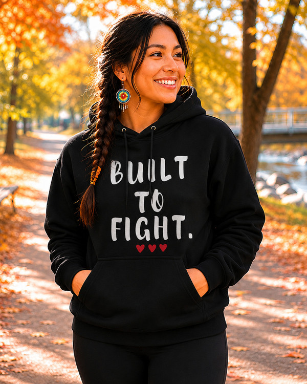 Smiling woman wearing a black hoodie featuring the inspirational quote “Built to Fight” in distressed white lettering with red heart accents, standing on a tree-lined path during autumn.