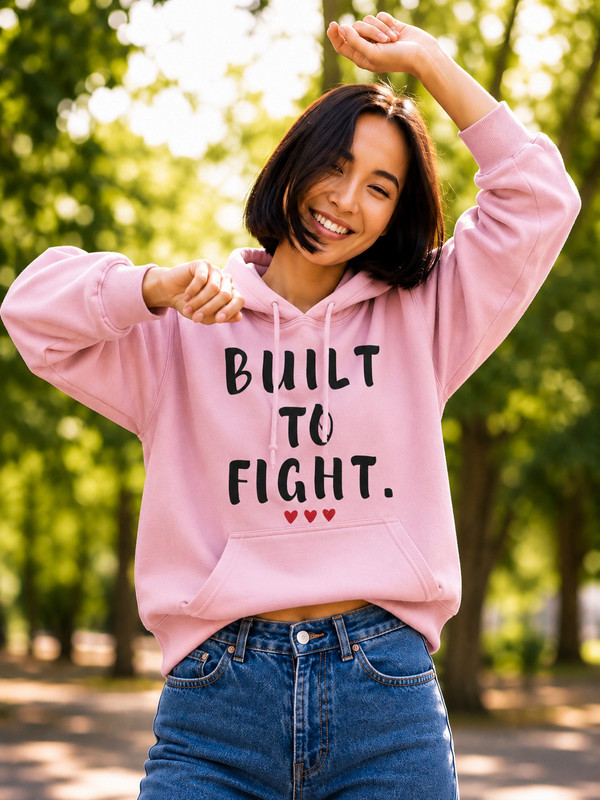 Smiling woman wearing a light pink hoodie featuring the inspirational quote “Built to Fight” in distressed black lettering with red heart accents, posing outdoors in a sunlit park setting.