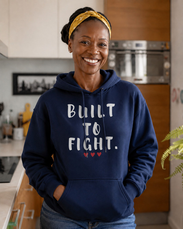 Smiling woman wearing a navy blue hoodie featuring the inspirational quote “Built to Fight” in distressed white lettering with red heart accents, standing in a bright kitchen setting.