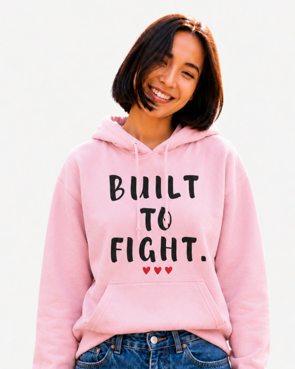 Smiling woman wearing a light pink hoodie featuring the inspirational quote “Built to Fight” in distressed black lettering with red heart accents, posing outdoors in a sunlit park setting.