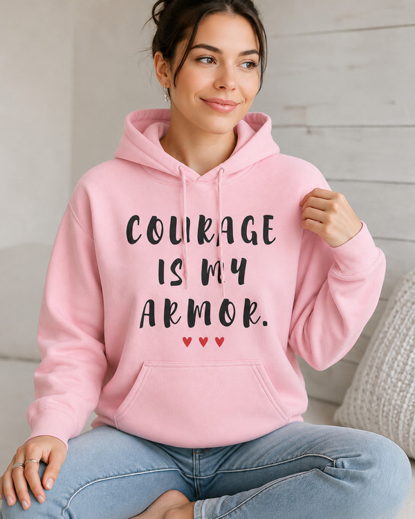Woman wearing a pink hoodie with the motivational quote “COURAGE IS MY ARMOR” and three red hearts, sitting indoors in a cozy setting, symbolizing strength, resilience, confidence, empowerment, and overcoming life’s challenges.