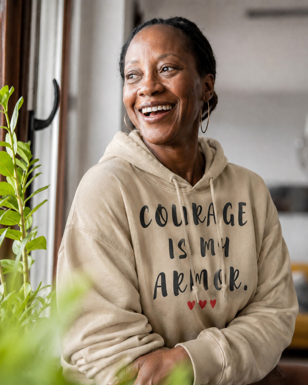 Woman wearing a sand-colored hoodie with the motivational quote “COURAGE IS MY ARMOR” and three red hearts, smiling by a window in a bright indoor setting, symbolizing strength, resilience, bravery, hope, empowerment, and overcoming life’s challenges.