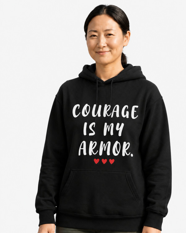 Woman wearing a black hoodie with the motivational quote “COURAGE IS MY ARMOR” and three red hearts, smiling in a workshop setting, symbolizing strength, resilience, determination, empowerment, and overcoming life’s challenges.