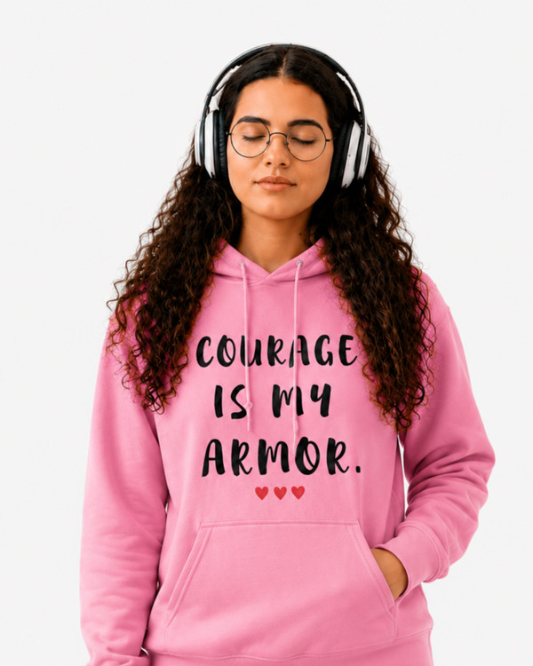 Woman wearing a pink hoodie with the motivational quote “COURAGE IS MY ARMOR” and three red hearts, sitting indoors in a cozy setting, symbolizing strength, resilience, confidence, empowerment, and overcoming life’s challenges.