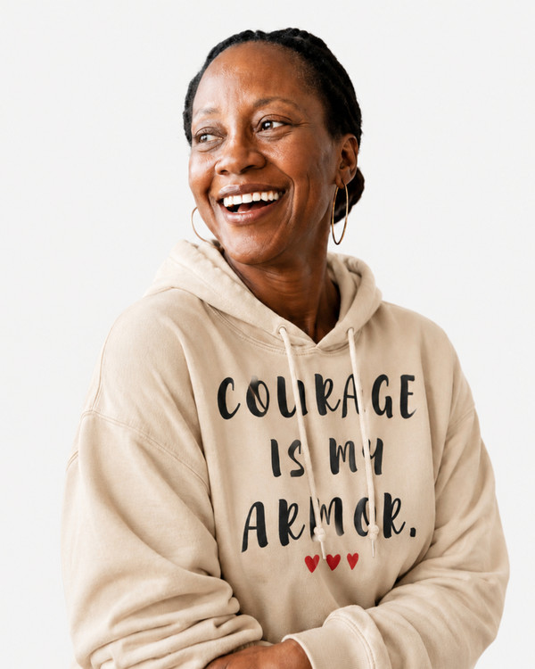 Woman wearing a sand-colored hoodie with the motivational quote “COURAGE IS MY ARMOR” and three red hearts, smiling by a window in a bright indoor setting, symbolizing strength, resilience, bravery, hope, empowerment, and overcoming life’s challenges.