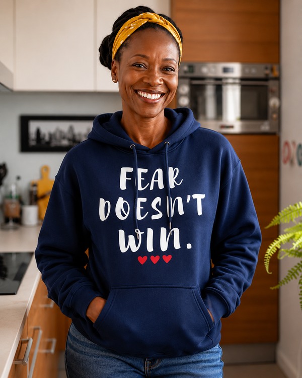 Woman wearing a navy blue hoodie with the motivational quote “FEAR DOESN’T WIN” and three red hearts, smiling indoors in a modern kitchen, symbolizing courage, resilience, hope, confidence, and overcoming life’s challenges.