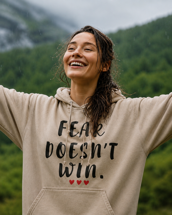 Woman wearing a sand-colored hoodie with the motivational quote “FEAR DOESN’T WIN” and three red hearts, standing outdoors with arms raised in a mountain landscape, symbolizing courage, resilience, hope, confidence, and overcoming fear.