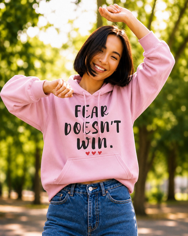 Woman wearing a pink hoodie with the motivational quote “FEAR DOESN’T WIN” and three red hearts, smiling and dancing outdoors in a sunlit park, symbolizing confidence, courage, positivity, resilience, and overcoming fear.