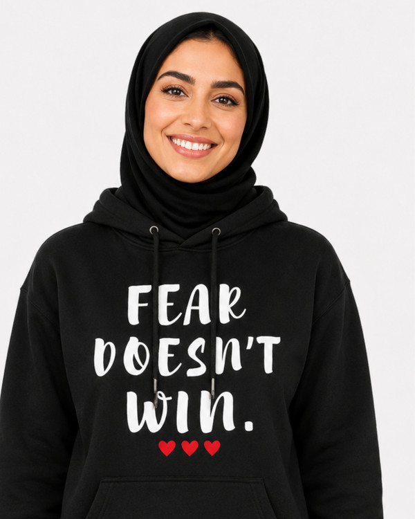 Stylish woman wearing a black hoodie with the inspirational quote “FEAR DOESN’T WIN” and three red hearts, walking outdoors at sunset, symbolizing courage, confidence, resilience, empowerment, and overcoming fear.