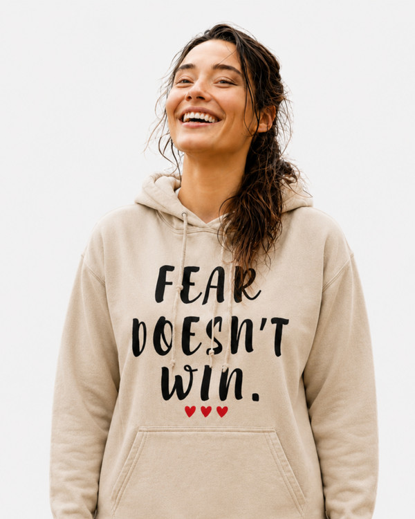 Woman wearing a sand-colored hoodie with the motivational quote “FEAR DOESN’T WIN” and three red hearts, standing outdoors with arms raised in a mountain landscape, symbolizing courage, resilience, hope, confidence, and overcoming fear.