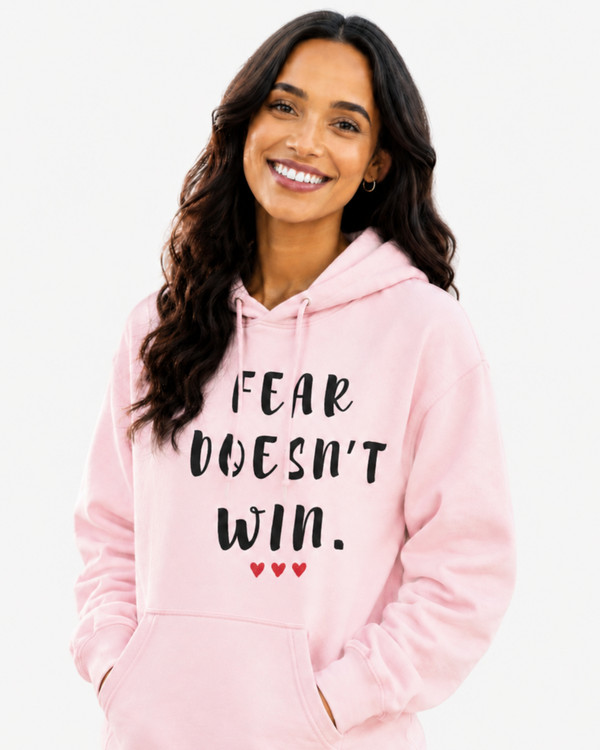 Woman wearing a pink hoodie with the motivational quote “FEAR DOESN’T WIN” and three red hearts, smiling and dancing outdoors in a sunlit park, symbolizing confidence, courage, positivity, resilience, and overcoming fear.