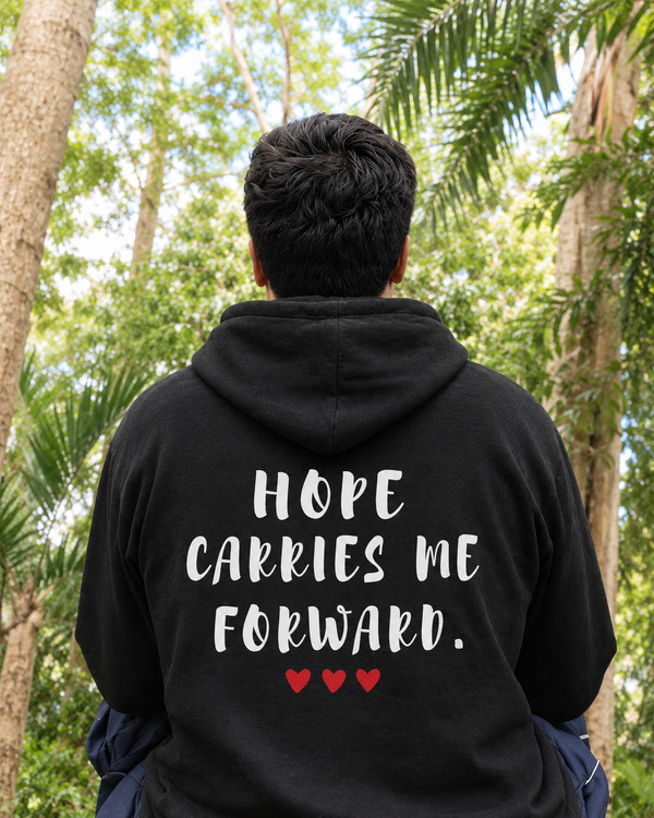 Person wearing a black hoodie outdoors in a tropical forest setting, viewed from the back, featuring the inspirational quote “Hope Carries Me Forward” in white lettering with red hearts beneath.