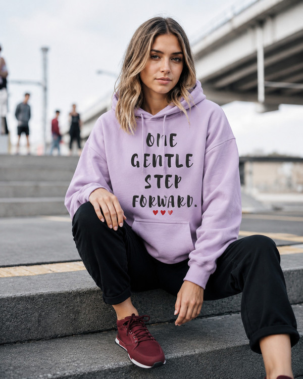 Young woman sitting on urban steps wearing a light pink hoodie featuring the inspirational quote “One Gentle Step Forward” in distressed black lettering with red heart accents.