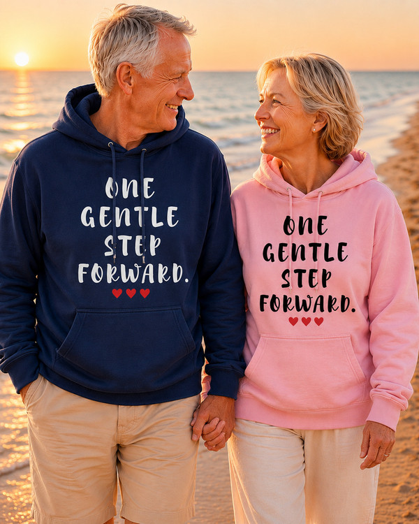 Smiling older couple walking hand in hand on a beach at sunset, wearing navy and pink hoodies featuring the inspirational quote “One Gentle Step Forward” with red heart accents.