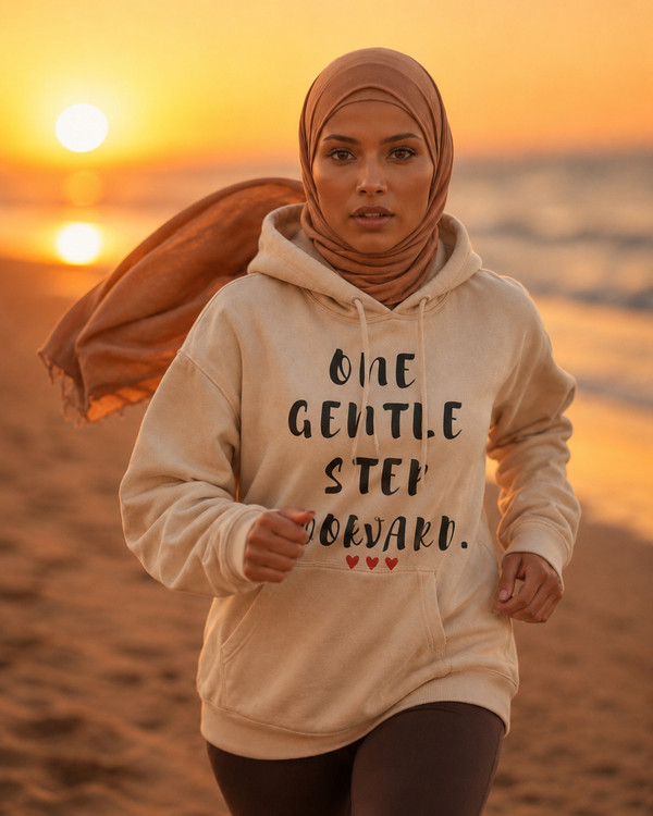 Woman running on a beach at sunset wearing a sand-colored hoodie featuring the inspirational quote “One Gentle Step Forward” in distressed black lettering with red heart accents.