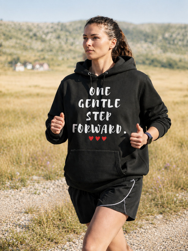 Woman jogging outdoors in a black hoodie featuring the inspirational quote “One Gentle Step Forward” in distressed white lettering, captured in a natural countryside setting.