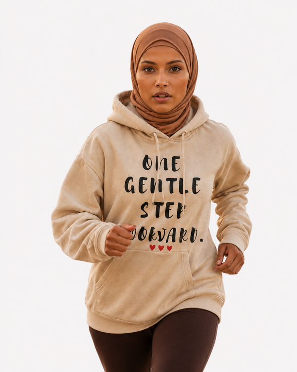 Woman running on a beach at sunset wearing a sand-colored hoodie featuring the inspirational quote “One Gentle Step Forward” in distressed black lettering with red heart accents.