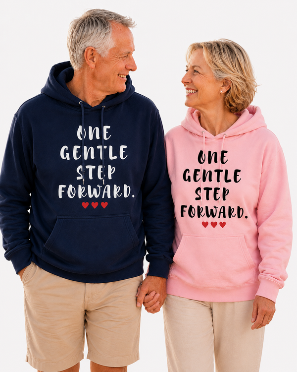 Smiling older couple walking hand in hand on a beach at sunset, wearing navy and pink hoodies featuring the inspirational quote “One Gentle Step Forward” with red heart accents.