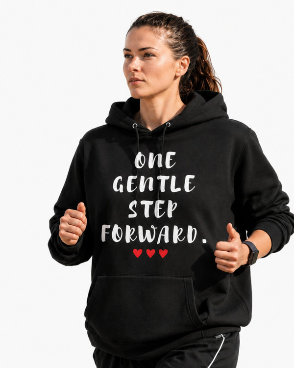 Woman jogging outdoors in a black hoodie featuring the inspirational quote “One Gentle Step Forward” in distressed white lettering, captured in a natural countryside setting.