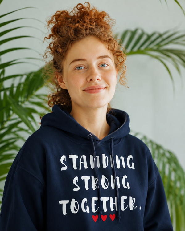 Smiling woman wearing a navy blue hoodie featuring the inspirational quote “Standing Strong Together” in white lettering with red heart accents, photographed indoors with lush green plants in the background.