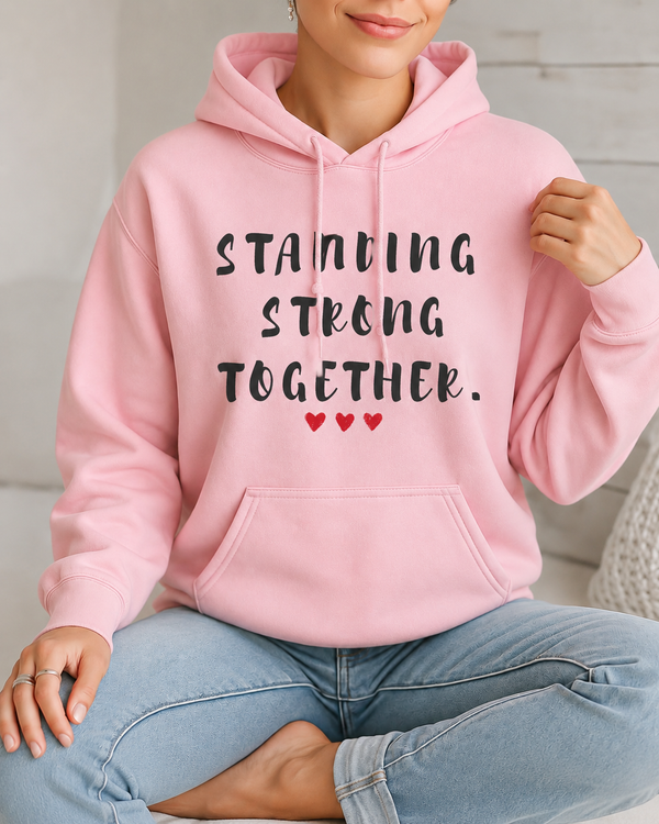 Woman wearing a light pink hoodie featuring the inspirational quote “Standing Strong Together” in distressed black lettering with red heart accents, seated indoors in a cozy lifestyle setting.