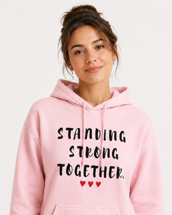 Woman wearing a light pink hoodie featuring the inspirational quote “Standing Strong Together” in distressed black lettering with red heart accents, seated indoors in a cozy lifestyle setting.