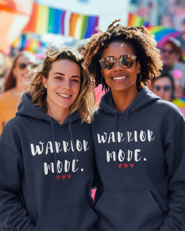 Two women wearing matching navy blue hoodies featuring the motivational quote “Warrior Mode” with heart accents, smiling at an outdoor community event, inspirational apparel celebrating strength, unity, and empowerment.