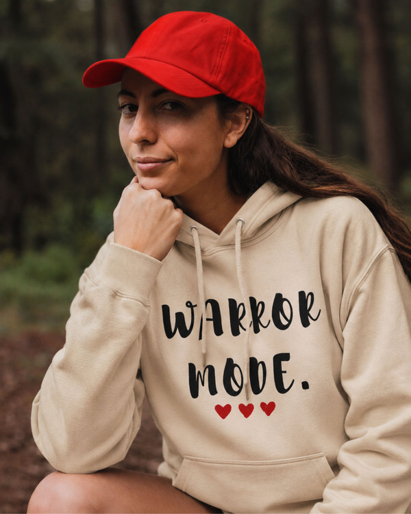 Woman wearing a beige hoodie featuring the motivational quote “Warrior Mode” with heart accents, paired with a red cap in an outdoor setting, inspirational apparel promoting strength, courage, and resilience.