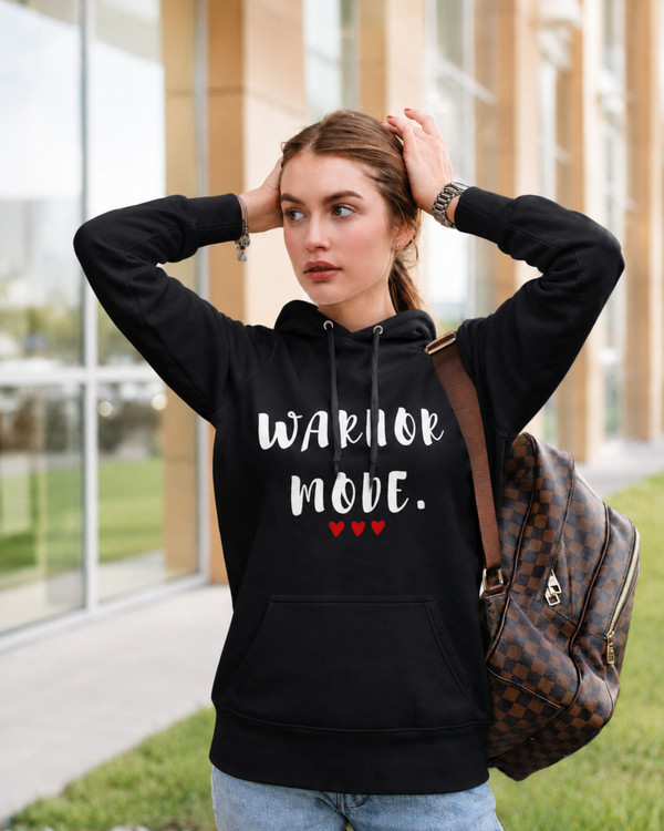 Woman wearing a black hoodie featuring the inspirational quote “Warrior Mode” with red heart accents, styled casually outdoors with a backpack, empowering motivational apparel for women.