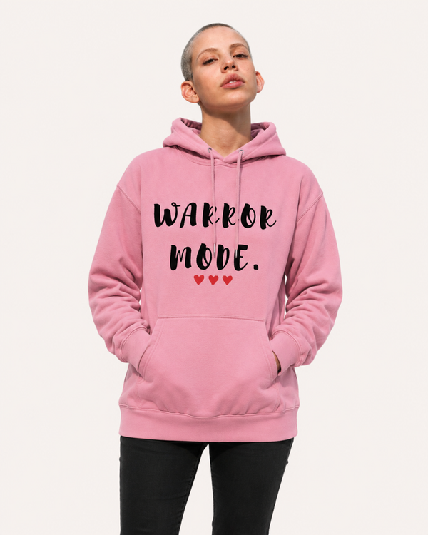 Woman wearing a pink hoodie with the motivational quote “Warrior Mode” and heart accents, seated on outdoor steps, stylish inspirational apparel promoting strength, resilience, and confidence.