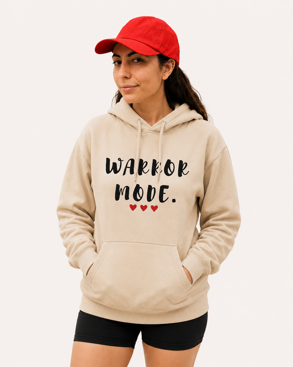 Woman wearing a beige hoodie featuring the motivational quote “Warrior Mode” with heart accents, paired with a red cap in an outdoor setting, inspirational apparel promoting strength, courage, and resilience.