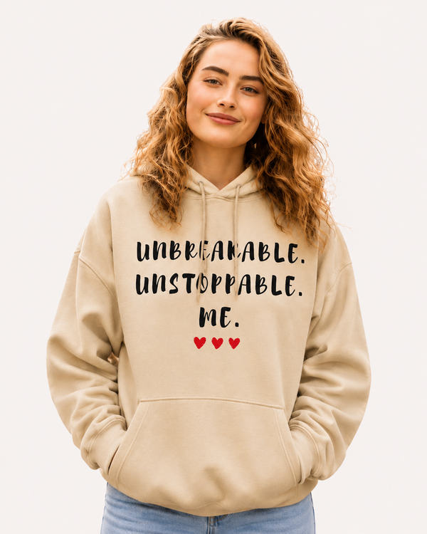 Woman wearing a beige hoodie featuring the empowering quote “Unbreakable. Unstoppable. Me.” with heart accents, inspirational apparel promoting confidence, resilience, strength, and self-empowerment in a casual outdoor setting.