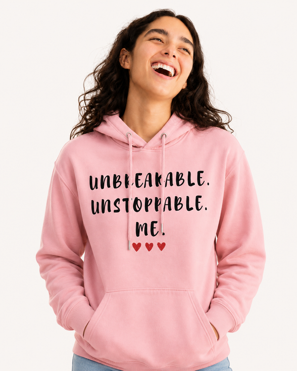 Woman wearing a pink hoodie featuring the empowering quote “Unbreakable. Unstoppable. Me.” with heart accents, smiling on a beach, inspirational apparel celebrating confidence, resilience, strength, and self-belief.