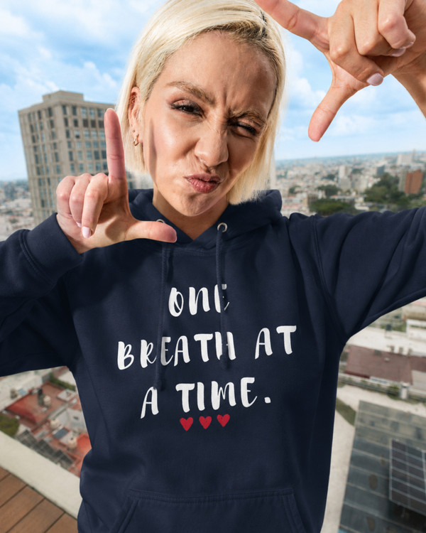 Woman wearing a navy blue hoodie featuring the inspirational quote “One Breath at a Time” with red heart accents, motivational apparel promoting mindfulness, resilience, self-care, and emotional well-being against a city skyline backdrop.