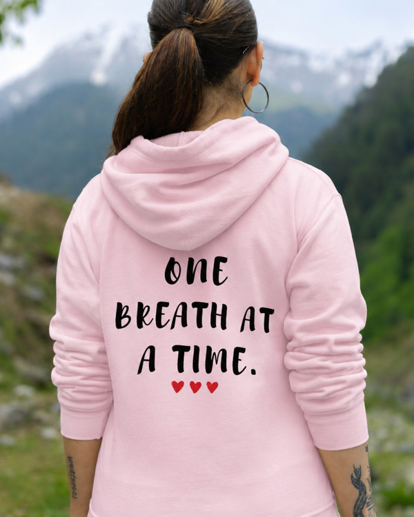 Back view of a woman wearing a pink hoodie featuring the inspirational quote “One Breath at a Time” with heart accents, motivational apparel promoting mindfulness, resilience, inner peace, and emotional strength in a mountain setting.