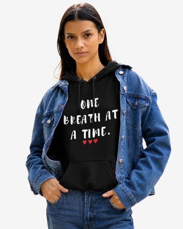 Woman wearing a black hoodie featuring the inspirational quote “One Breath at a Time” with red heart accents, layered under a denim jacket, motivational apparel promoting mindfulness, resilience, hope, and inner strength.