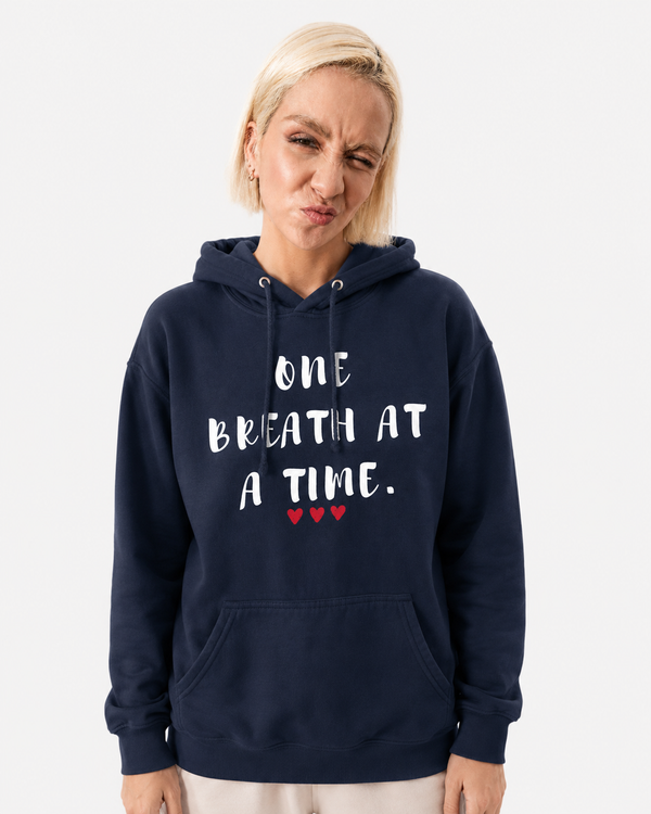 Woman wearing a navy blue hoodie featuring the inspirational quote “One Breath at a Time” with red heart accents, motivational apparel promoting mindfulness, resilience, self-care, and emotional well-being against a city skyline backdrop.