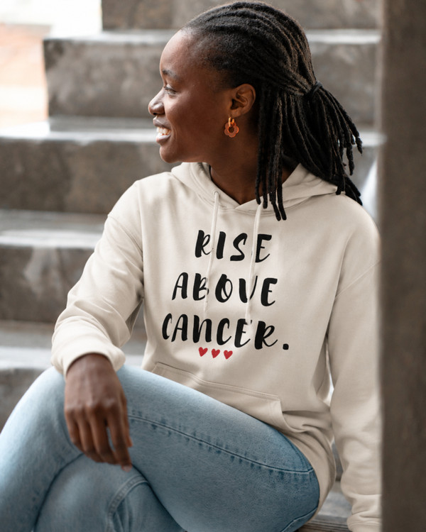 Woman wearing a beige hoodie featuring the inspirational quote “Rise Above Cancer” with red heart accents, seated on outdoor steps, motivational apparel promoting hope, courage, resilience, strength, and cancer awareness.