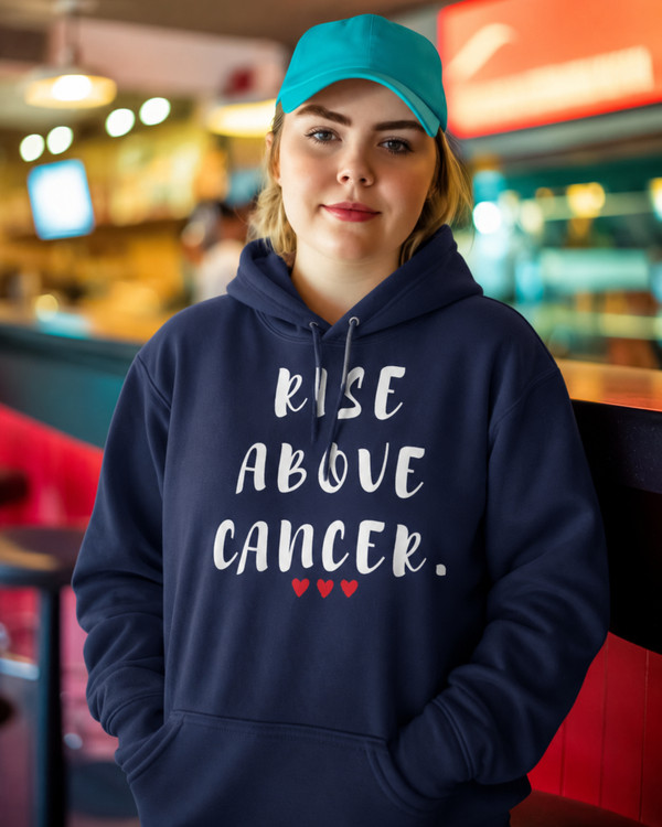 Woman wearing a navy blue hoodie featuring the inspirational quote “Rise Above Cancer” with red heart accents, motivational apparel promoting strength, hope, courage, and cancer awareness in a casual setting.