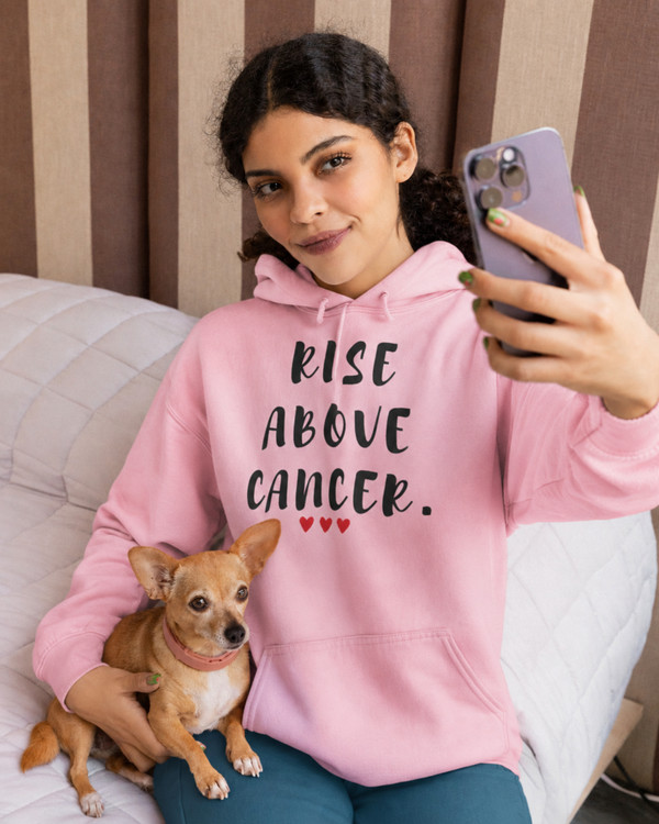 Woman wearing a pink hoodie featuring the inspirational quote “Rise Above Cancer” with red heart accents, taking a selfie while holding a small dog, motivational apparel promoting hope, strength, resilience, and cancer awareness.