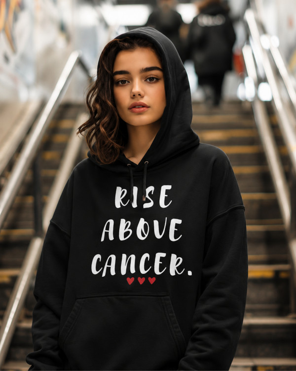 Woman wearing a black hoodie featuring the inspirational quote “Rise Above Cancer” with red heart accents, standing on a staircase, motivational apparel promoting hope, courage, resilience, strength, and cancer awareness.